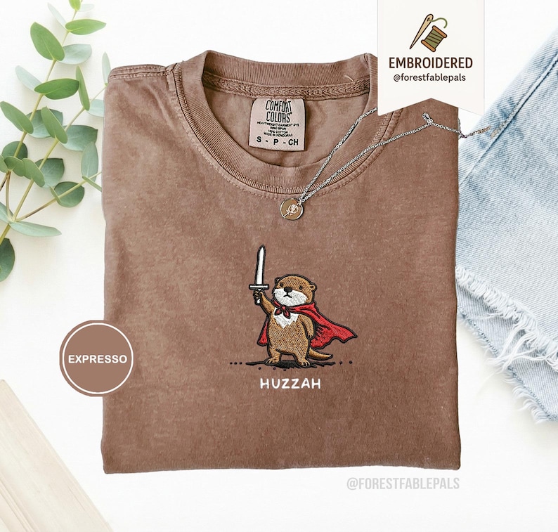 May include: Espresso-colored t-shirt with an embroidered otter design. The otter, wearing a red cape and holding a sword, is embroidered above the word "Huzzah". The shirt has a crew neck and short sleeves. A silver necklace with a pendant is also visible.