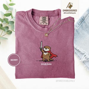 May include: Berry-colored t-shirt with an embroidered otter design. The otter is depicted holding a sword and wearing a red cape. The word "Huzzah" is embroidered below the otter. A gold necklace is draped on the shirt.