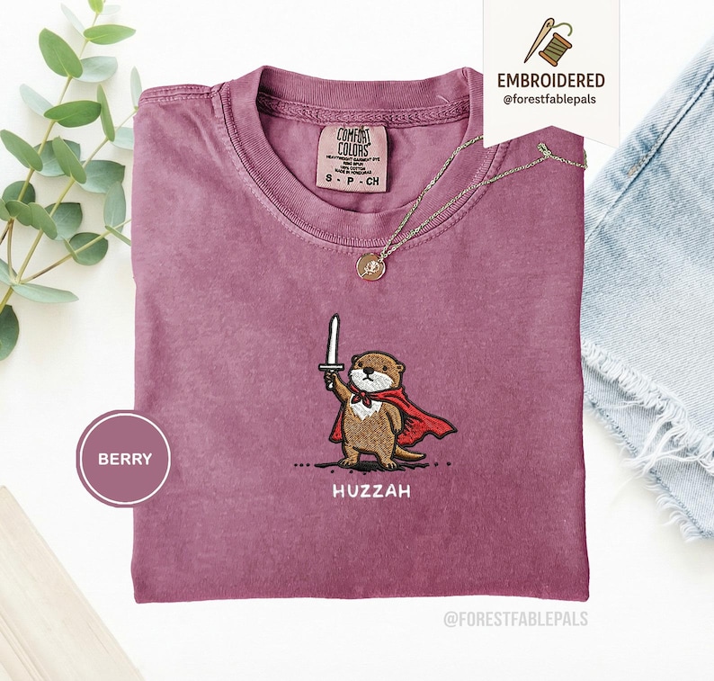 May include: Berry-colored t-shirt with an embroidered otter design. The otter is depicted holding a sword and wearing a red cape. The word "Huzzah" is embroidered below the otter. A gold necklace is draped on the shirt.