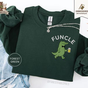 以下が含まれることがあります： 「FUNCLE」の文字が白で刺繍されたフォレストグリーンのスウェットシャツ。蝶ネクタイとビールを持った漫画のアリゲーターが描かれています。丸いペンダントが付いたシルバーのネックレスがスウェットシャツの上にかけられています。