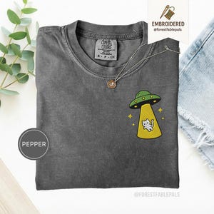 Op de afbeelding: Een antracietgrijs T-shirt met een geborduurd ontwerp van een groene UFO met een gele tractorstraal die een kleine witte kat optilt. Het shirt heeft een ronde hals en een Comfort Colors label in de kraag. De tekst PEPPER is zichtbaar.