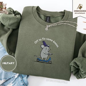May include: Olive green sweatshirt with the embroidered phrase "Off to fix some knees" and a cartoon mouse riding a blue scooter. The sweatshirt has a crew neck and long sleeves. A silver necklace with a pendant is draped across the top.