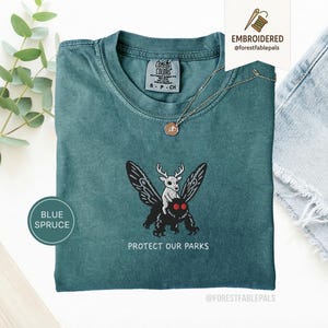 Pode incluir: Camiseta azul abeto com um design bordado com um veado montado numa traça com o texto "PROTECT OUR PARKS". A camisa tem gola redonda e mangas curtas. Um colar de ouro com um pingente também é visível.