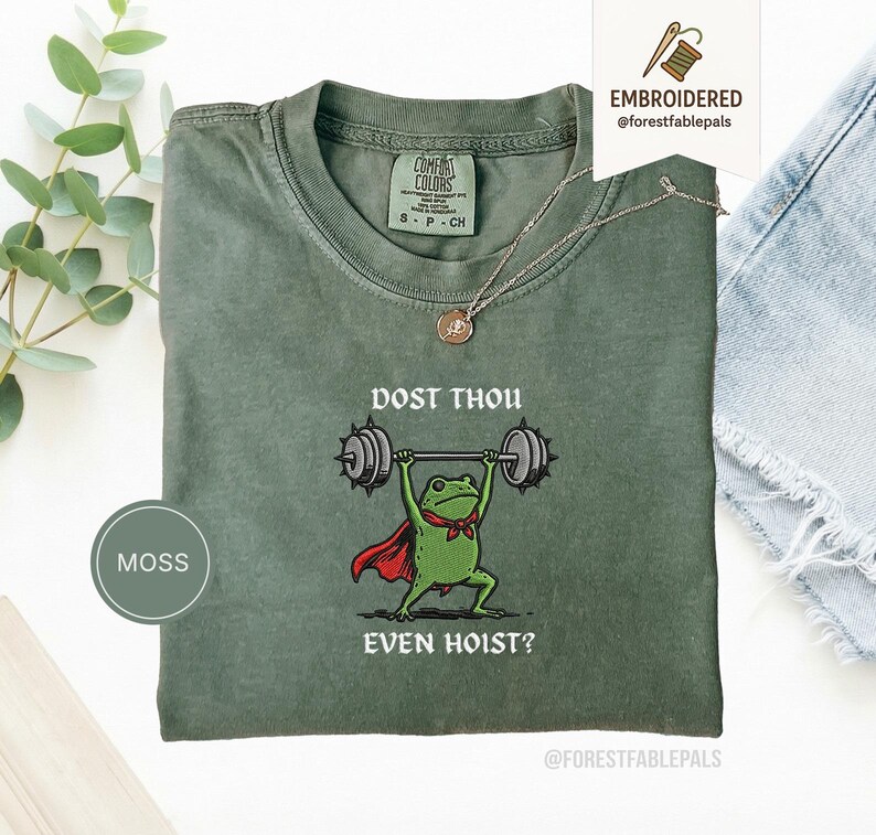 May include: Moss green t-shirt featuring a frog lifting weights, wearing a red cape and bow tie. The text reads "DOST THOU EVEN HOIST?" The shirt has a crew neck and short sleeves. A silver necklace is draped over the shirt. The shirt is a size S-P-CH.