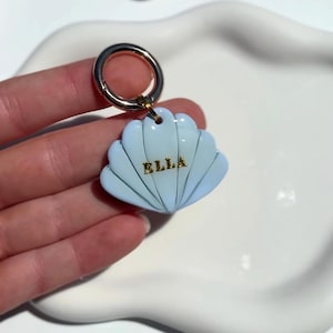 May include: A light blue seashell-shaped keychain with the name "ELLA" in gold lettering. The keychain has a gold-colored ring and clasp. The seashell design has a textured surface and a gradient effect.