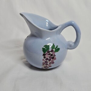 May include: A light blue ceramic pitcher with a rounded body and a curved handle. The exterior features a hand-painted design of a cluster of purple grapes with green leaves. The pitcher has a wide pouring spout and a smooth, glossy finish.