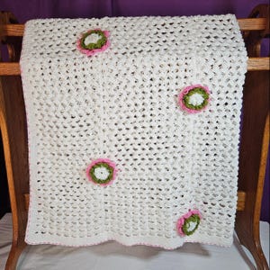 May include: A white crocheted blanket with a floral design. The blanket has a lacy pattern and is adorned with several pink and green flower appliqués. The blanket is draped over a wooden rack, showcasing its texture and design.