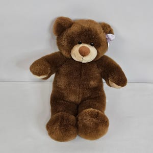 May include: A plush teddy bear with dark brown fur, a lighter tan muzzle, and a brown nose. The bear features small black eyes and a light lavender ribbon tied behind its left ear. The paws are a lighter tan color, matching the muzzle.