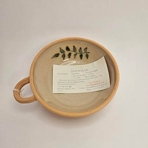 May include: A handmade ceramic egg baker with a handle, featuring a light brown exterior and a beige interior. The rim and handle are a darker brown. The interior has a green leaf design. A recipe card is inside the bowl.
