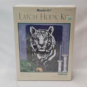 May include: A "WonderArt" latch hook kit featuring a white tiger design. The kit includes the materials to create a tapestry with an approximate size of 24" x 34". The box also includes the text "TAPISSERIE AU CROCHET GANCHILLO DE LENGÜETA".