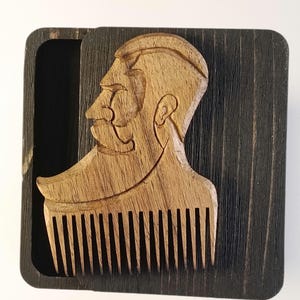 Puede incluir: Peine de barba de madera con un perfil tallado de un hombre con barba y bigote. El peine es de color marrón claro y descansa en una caja de madera marrón oscuro. Los dientes del peine son visibles y la caja tiene una tapa corredera.