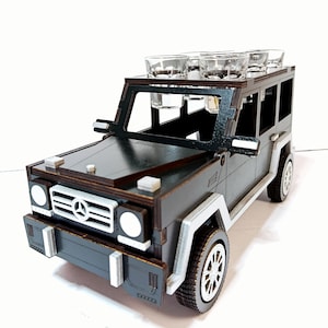 May include: A wooden model of a dark gray SUV with clear shot glasses on top. The model has white accents on the wheels and bumpers. The SUV has a detailed design, including headlights and a front grill.