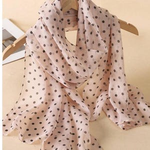 May include: A light pink scarf with a polka dot pattern. The scarf is draped over a wooden hanger. The polka dots are dark gray and evenly spaced across the fabric. The scarf appears to be made of a lightweight material.