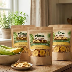 May include: Three brown paper bags of Adom Golden Plantain chips with a clear window showing the chips. A bowl of green plantains and a small plate of chips are in front of the bags. The bags have a green and yellow label.