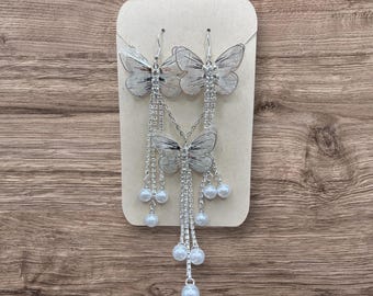 May include: A silver-colored butterfly jewelry set, including a necklace and earrings, displayed on a neutral-toned card. The butterfly pendants and earrings have intricate wing details and are adorned with dangling chains and faux pearls.