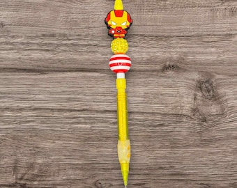 May include: A yellow mechanical pencil with a decorative top featuring an Iron Man character. The pencil has a series of beads in yellow, red, and white. The Iron Man figure is red, yellow, and black.