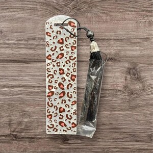 Puede incluir: Marcapáginas rectangular con diseño de estampado de leopardo en marrón y rosa sobre fondo blanco. Tiene una borla negra sujeta con un cordón negro. La borla está en una funda de plástico transparente.