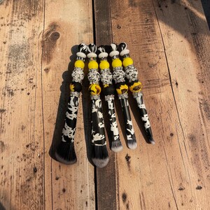May include: Five makeup brushes with black and white cow print handles and decorative beaded accents. The beads feature yellow, black, and white patterns, including sunflower designs, interspersed with silver-toned spacers and rings.