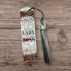 May include: A decorative bookmark with a leopard print and the text "Act like a lady, think like a boss" in black and pink. The bookmark has a black tassel attached to the top with a black string.