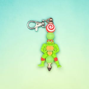 Könnte beinhalten: Ein grüner Teenage Mutant Ninja Turtle-Anhänger mit roter Maske, orangefarbenem Panzer und roten Knieschonern. Der Anhänger ist an einem silberfarbenen Verschluss mit einer rot-weißen Wirbelperle und grünen Perlen befestigt.