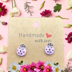 以下が含まれることがあります： 「Handmade with love」の文字が入ったベージュ色のカードに、てんとう虫のスタッドピアス。ピアスは薄いピンク色で黒い水玉模様、銀色のトップが付いています。背景はピンク色で花が飾られています。