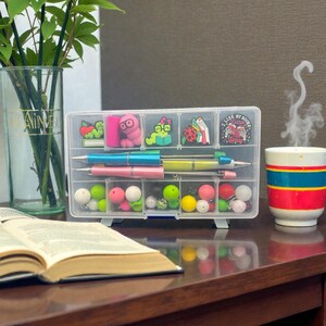 May include: A clear plastic organiser filled with pens, decorative charms, and colourful beads. The organiser sits on a desk next to a cup with blue, red, and yellow stripes and an open book.