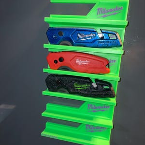 May include: A bright green plastic wall-mounted organizer rack featuring the Milwaukee logo in purple. The rack holds three Milwaukee Fastback utility knives in blue, red, and black, with two empty slots remaining at the bottom of the unit.