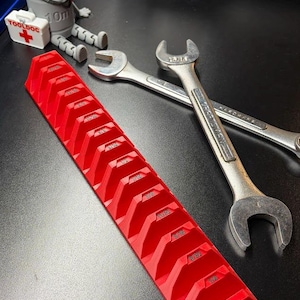 May include: A red wrench organizer with labeled slots, next to two silver wrenches. A gray tool-themed figurine and a white toolbox with the text "TOOLDOC" are also present. The tools are on a black surface.
