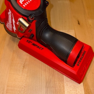 May include: A red Milwaukee Fuel Brushless 1/2 inch impact wrench resting in a red plastic ToolDoc mount. The tool features a black textured grip, a metal belt clip, and clear branding on the housing. The mount is labeled 1/2 inch impact.