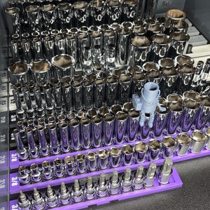 May include: A collection of shiny chrome sockets and bits, neatly organized in a gray toolbox. The sockets are arranged in rows, with a purple tray holding smaller bits. A small, gray 3D-printed object sits among the tools.