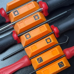 May include: An assortment of tools, including screwdrivers and picks, are displayed. The screwdrivers have black handles with red accents and orange holders. The orange holders have a square design with a symbol in the center.