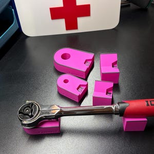 May include: A set of pink tool holders and a ratchet wrench. The tool holders are in various shapes and sizes, designed to cradle the wrench. The background includes a white sign with the text "TOOL DOC" and a red cross.