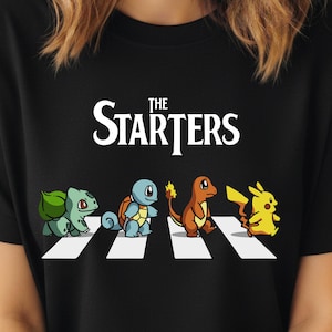 May include: Black t-shirt featuring a parody of the Beatles' Abbey Road album cover, with the text "The Starters" above four cartoon Pokémon characters crossing a crosswalk. The characters include Bulbasaur, Squirtle, Charmander, and Pikachu.