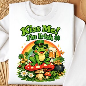 Puede incluir: Sudadera blanca con un diseño del Día de San Patricio. El diseño presenta una rana verde con sombrero, sosteniendo una cerveza y sentada en una seta. El texto "Kiss Me! I'm Irish" está encima de la rana, con un arcoíris y tréboles.