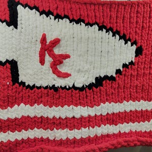 Pode incluir: Um cobertor de malha vermelho e branco com uma grande forma de ponta de flecha branca no centro. A ponta da flecha é delineada em preto e apresenta um logotipo "KC" vermelho. O cobertor tem listras brancas na parte inferior.