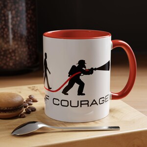 May include: White ceramic mug with a red interior and handle. The mug features a black silhouette of a firefighter with a hose and the words "OF COURAGE". A spoon and a macaron are in the foreground.
