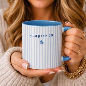 Könnte beinhalten: Eine weiße Keramiktasse mit blauen Streifen und blauem Innenraum. Die Tasse hat einen blauen Henkel und trägt den Text "chapter 18" mit einer kleinen floralen Illustration. Die Tasse wird von einer Person gehalten, die einen cremefarbenen Pullover trägt.