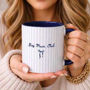 May include: White ceramic mug with blue vertical stripes and a blue interior. The mug has the text "Boy Mum Club" in blue script with a bow graphic. The handle and rim are navy blue.