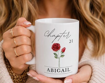 May include: White ceramic mug with the text "Chapter 21" and the name "Abigail" below a red rose illustration. The mug has a handle and is held by a person.
