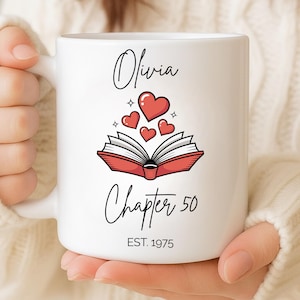 以下が含まれることがあります： 白いセラミックマグカップ。名前「Olivia」が筆記体で書かれ、開いた本とハートのグラフィック、テキスト「Chapter 50」と「EST. 1975」が描かれています。 マグカップは人が持っています。