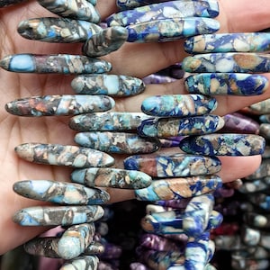 Può includere: Primo piano di una collezione di perline oblunghe nei toni del blu, marrone e crema. Le perline hanno un aspetto maculato e marmorizzato, suggerendo che siano fatte di pietra o di un materiale simile. Le perline sono infilate, pronte per la creazione di gioielli.