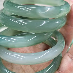 May include: A collection of jade bangles in varying shades of green. The smooth, polished surfaces reflect light, highlighting the natural variations in color. The bangles are stacked, showcasing their circular design and translucent quality.
