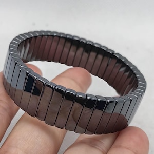 Peut inclure: Un bracelet gris foncé composé de segments rectangulaires et polis. Le bracelet est flexible et semble être fait d'un matériau métallique. Les segments sont reliés pour former une boucle continue.