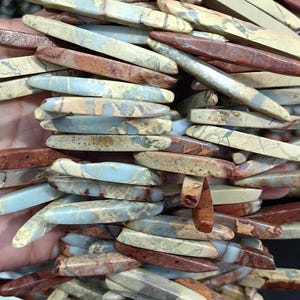 Puede incluir: Primer plano de una colección de cuentas de piedras preciosas rectangulares y alargadas en tonos rojos, beige y azul grisáceo. Las cuentas tienen un acabado pulido y están ensartadas, mostrando variaciones naturales de color y patrón. Ideal para bisutería.