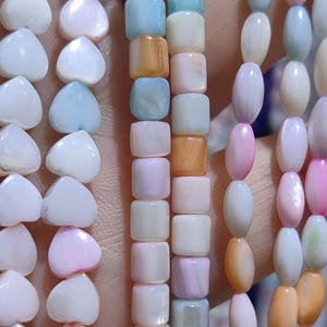 Pode incluir: Close-up de vários fios de contas em várias formas e cores. As contas incluem desenhos em forma de coração, quadrados e ovais em tons de branco, rosa, azul e laranja. As contas são enfiadas, criando uma exibição colorida.