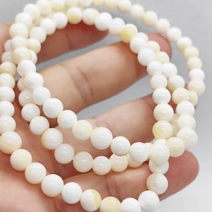 Könnte beinhalten: Drei Stränge weißer Perlenarmbänder mit einem cremefarbenen, cremeweißen Farbton. Die Perlen sind rund und glatt und erzeugen einen zarten und eleganten Look. Die Armbänder werden in einer Hand gehalten.