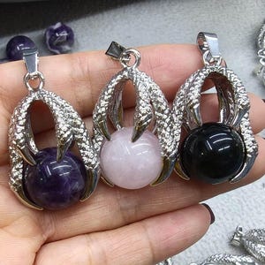 Puede incluir: Tres colgantes de piedras preciosas con un diseño de garra que sostiene una esfera. Las esferas son moradas, rosas y negras. Los colgantes tienen un acabado plateado y se muestran sobre un fondo claro.