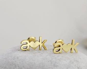 May include: A pair of gold-coloured stud earrings. Each earring features the letters "a" and "k" with a heart symbol between them. The earrings are displayed on a light grey, textured surface.