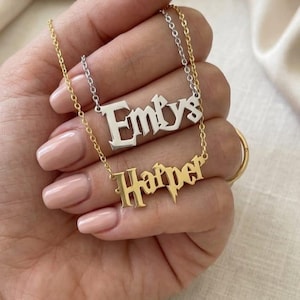 May include: Two name necklaces featuring a gothic-style font. One necklace is silver-toned with the name Emlys, and the other is gold-toned with the name Harper, which includes a lightning bolt detail on the letter p. Both have thin chains.