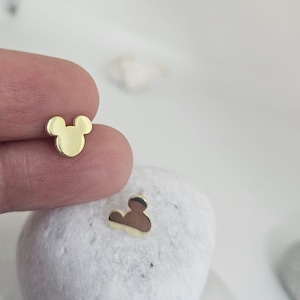 May include: Gold-colored stud earrings in the shape of a mouse head. One earring is held between fingers, while the other rests on a white stone. The earrings have a smooth, polished finish and a classic design.